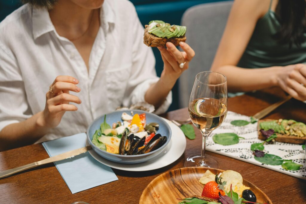 A colorful dining scene featuring avocado toast, seafood, and white wine shared among friends.